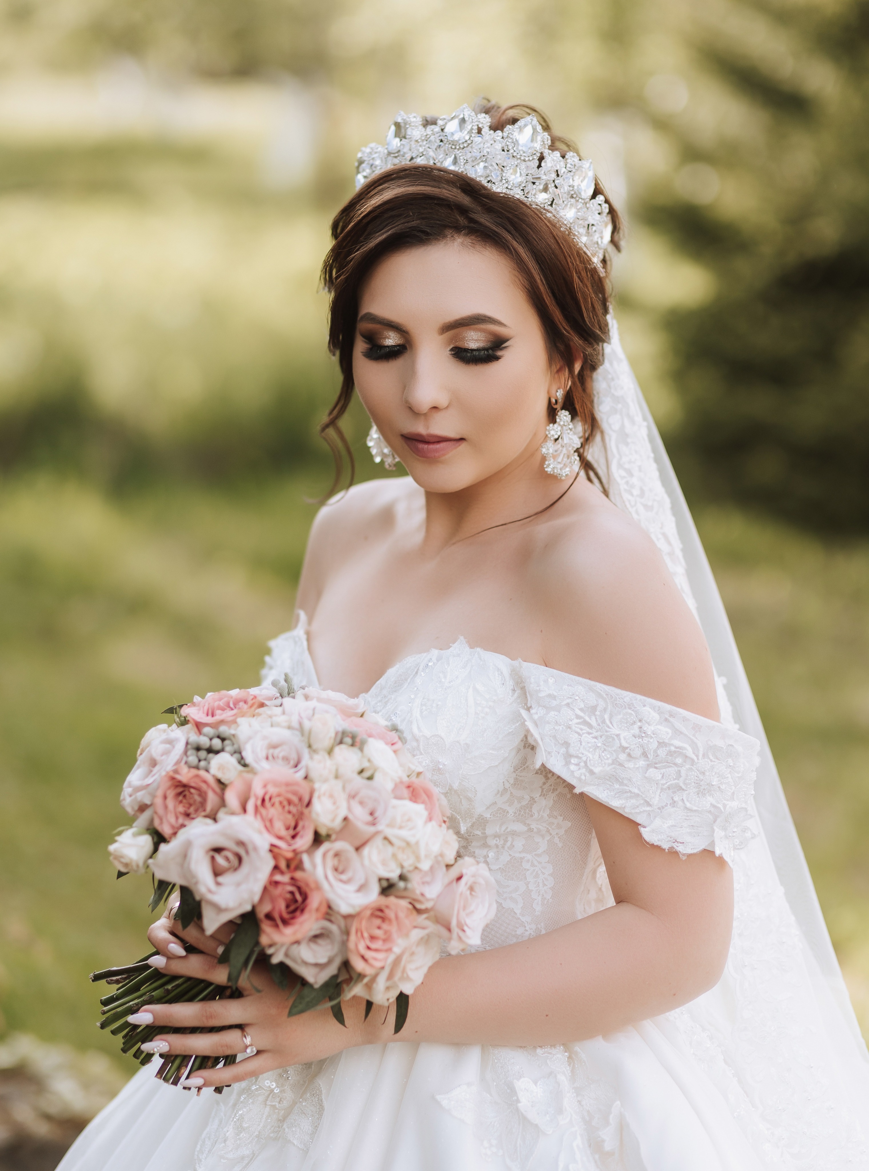 Bridal portrait of a bride in an off-the-shoulder lace gown and veil, holding a blush rose bouquet against soft green foliage
