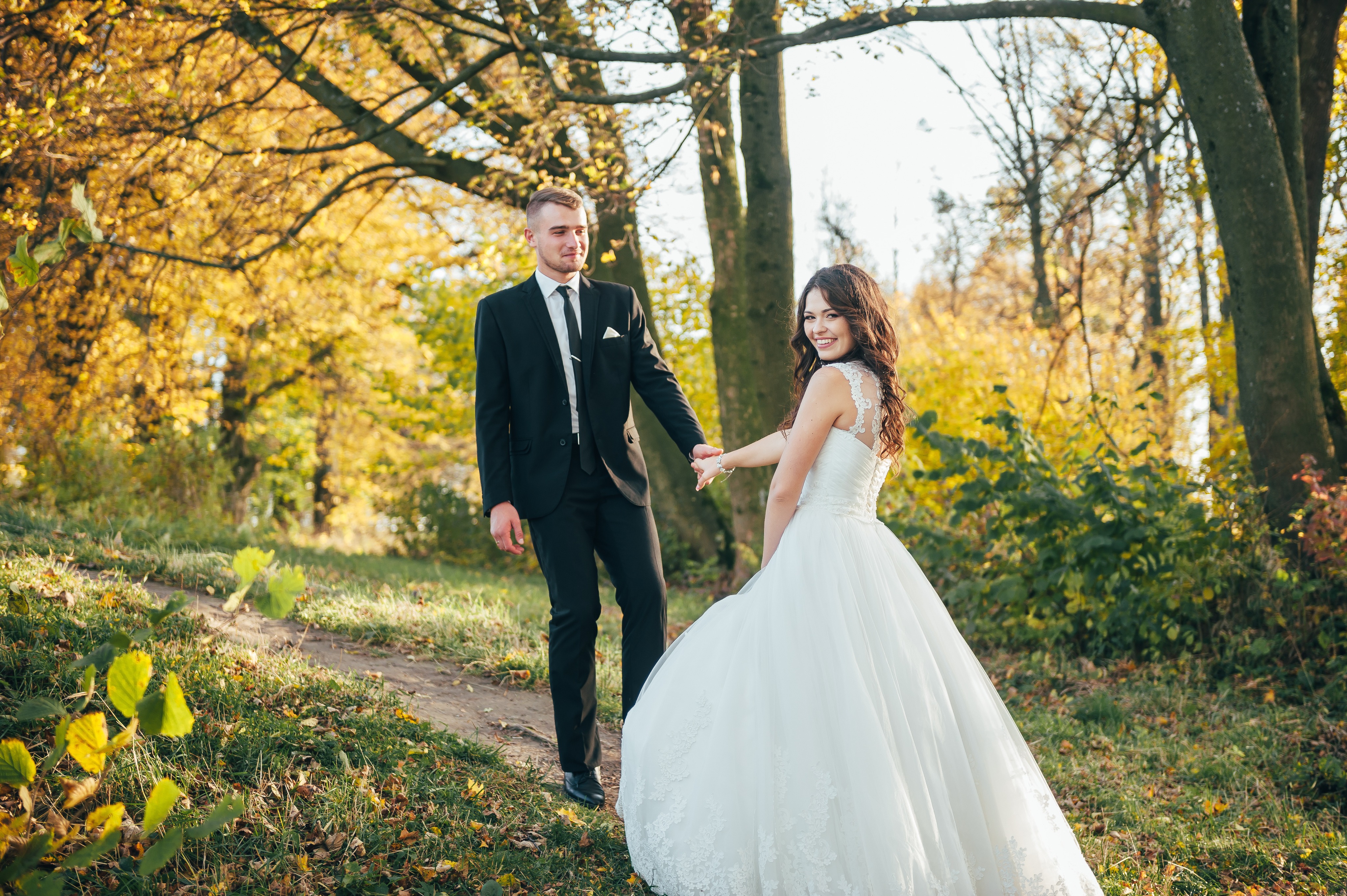A couple holding hands on a woodland path in golden autumn light, the bride looking back and smiling