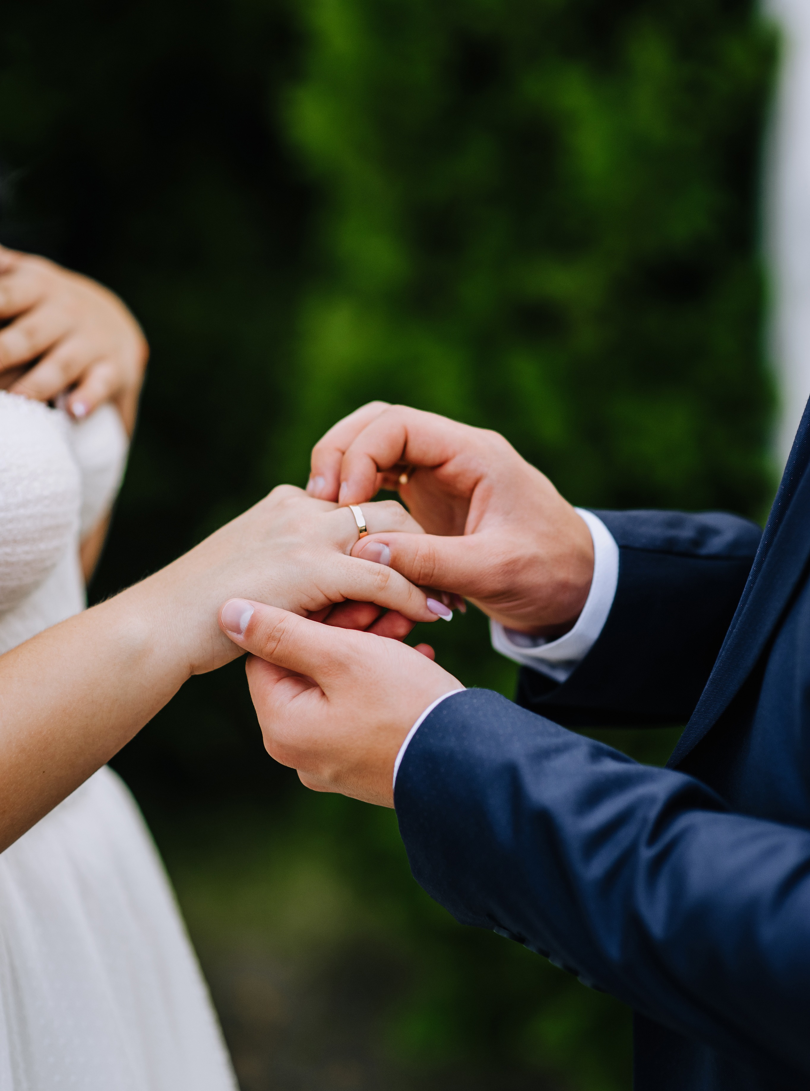 Close-up of a groom in a navy suit placing a gold wedding band on the bride's finger
