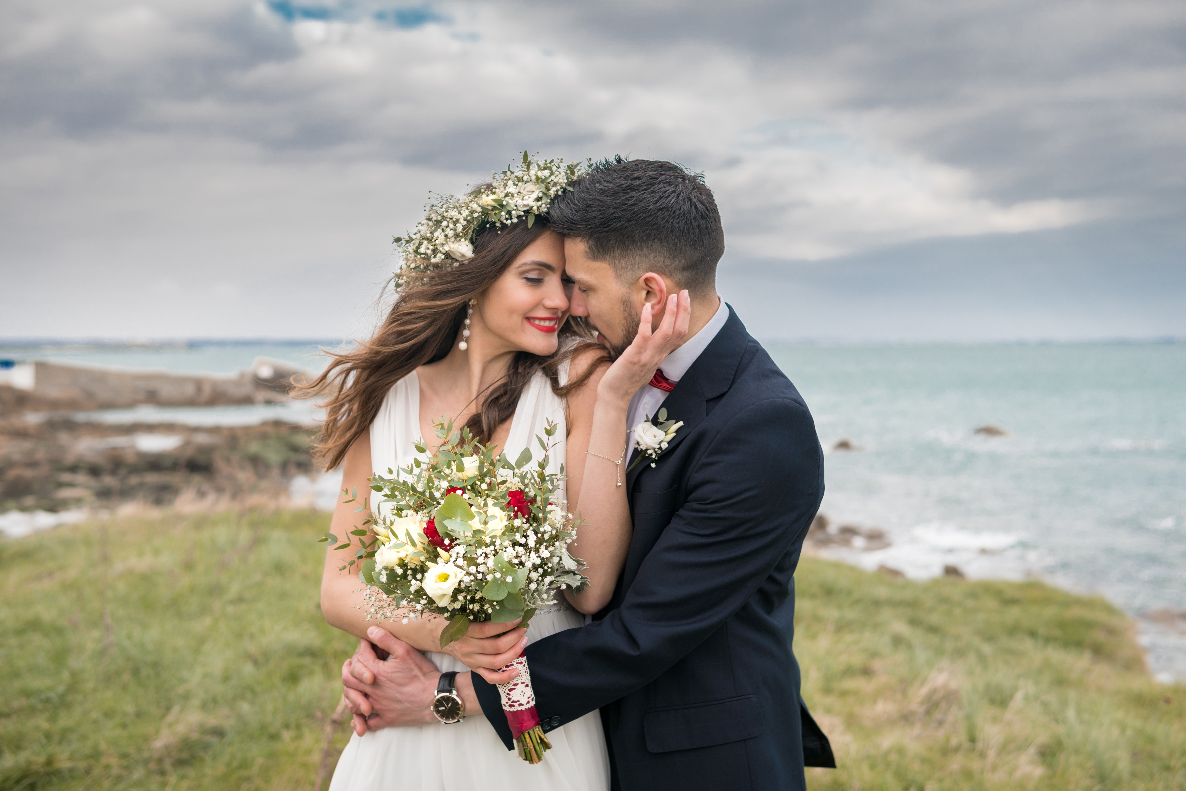 A couple embracing on a grassy clifftop above the sea, the bride wearing a flower crown and holding a bouquet